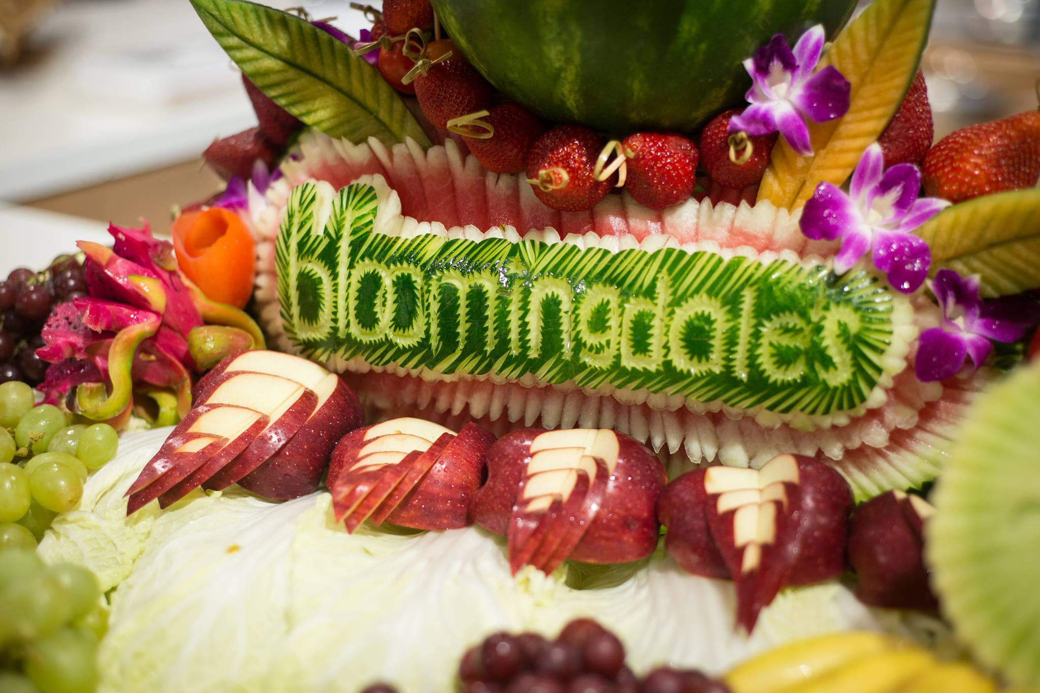 A watermelon carved with the Bloomingdale's wordmark, set in a fruit display with strawberries, orchids, dragon fruit and fanned apples.