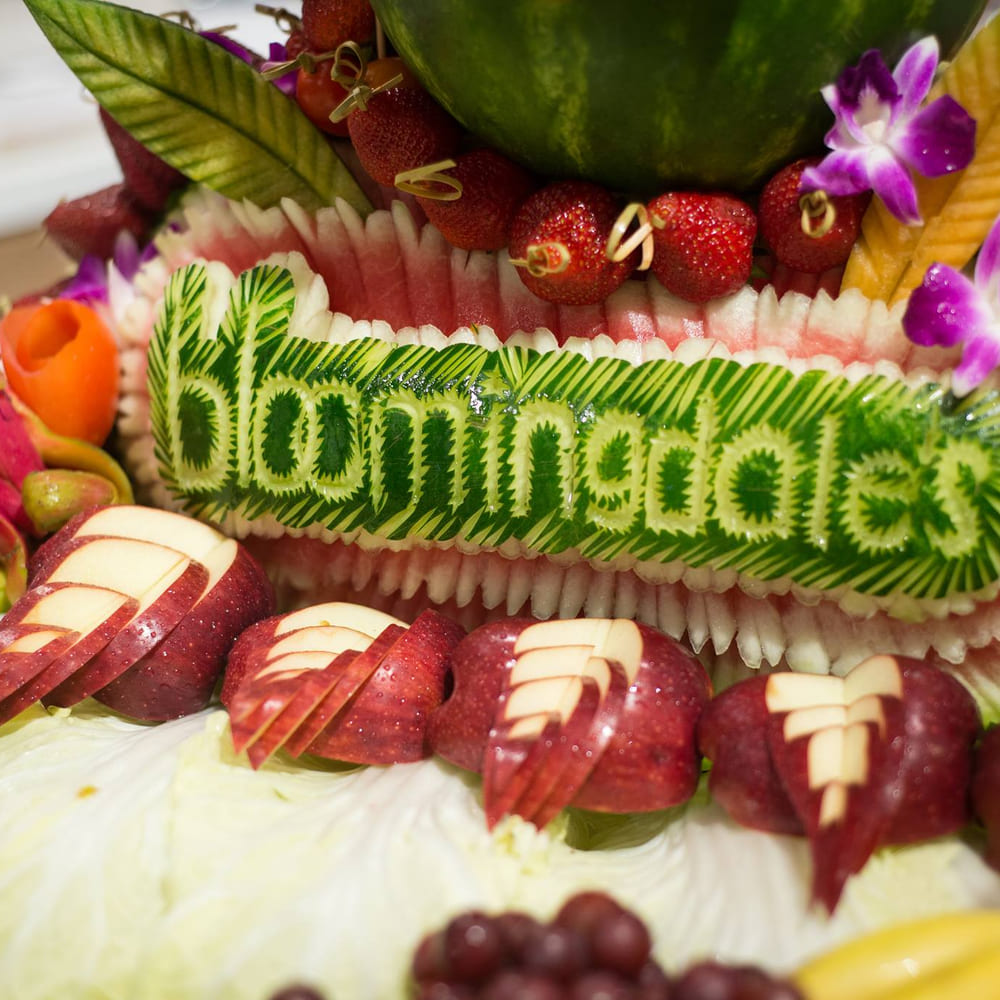 A watermelon carved with the Bloomingdale's wordmark, set in a fruit display with strawberries, orchids, dragon fruit and fanned apples.