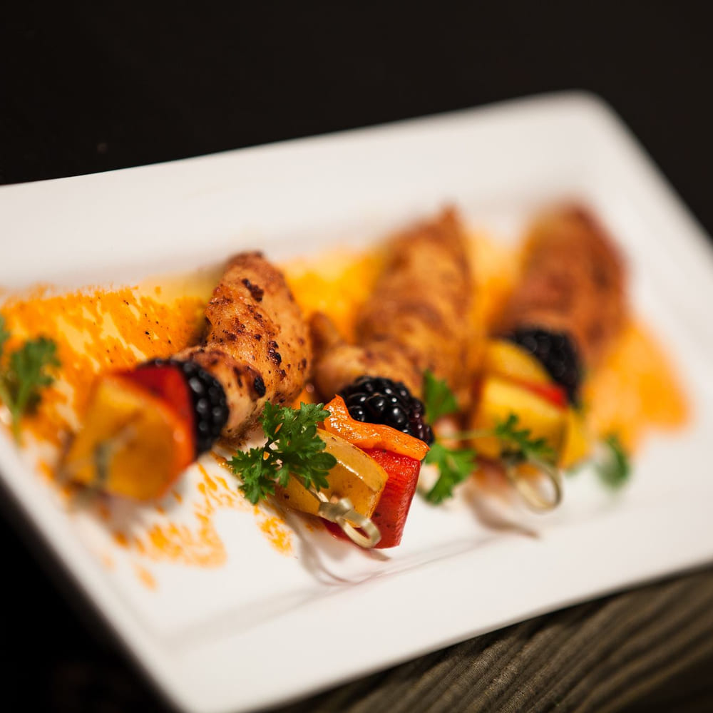 Grilled chicken skewers with blackberries, peppers and parsley on a white rectangular plate.
