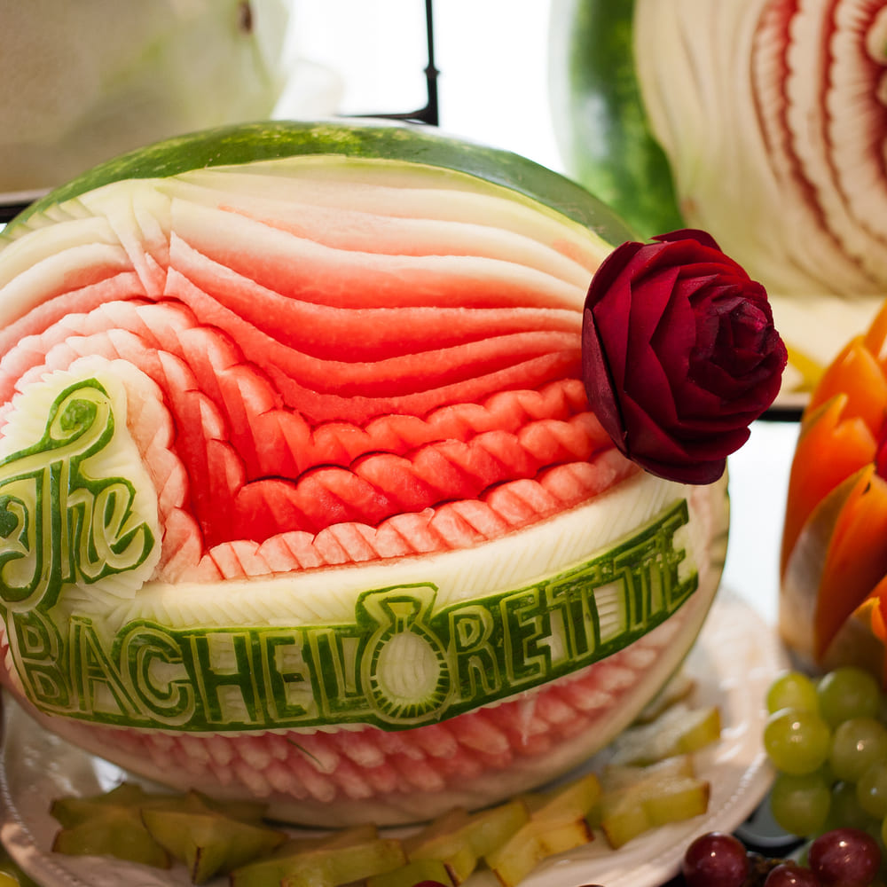 A watermelon carved with the words The Bachelorette and a rose, set among carved melon and grapes.