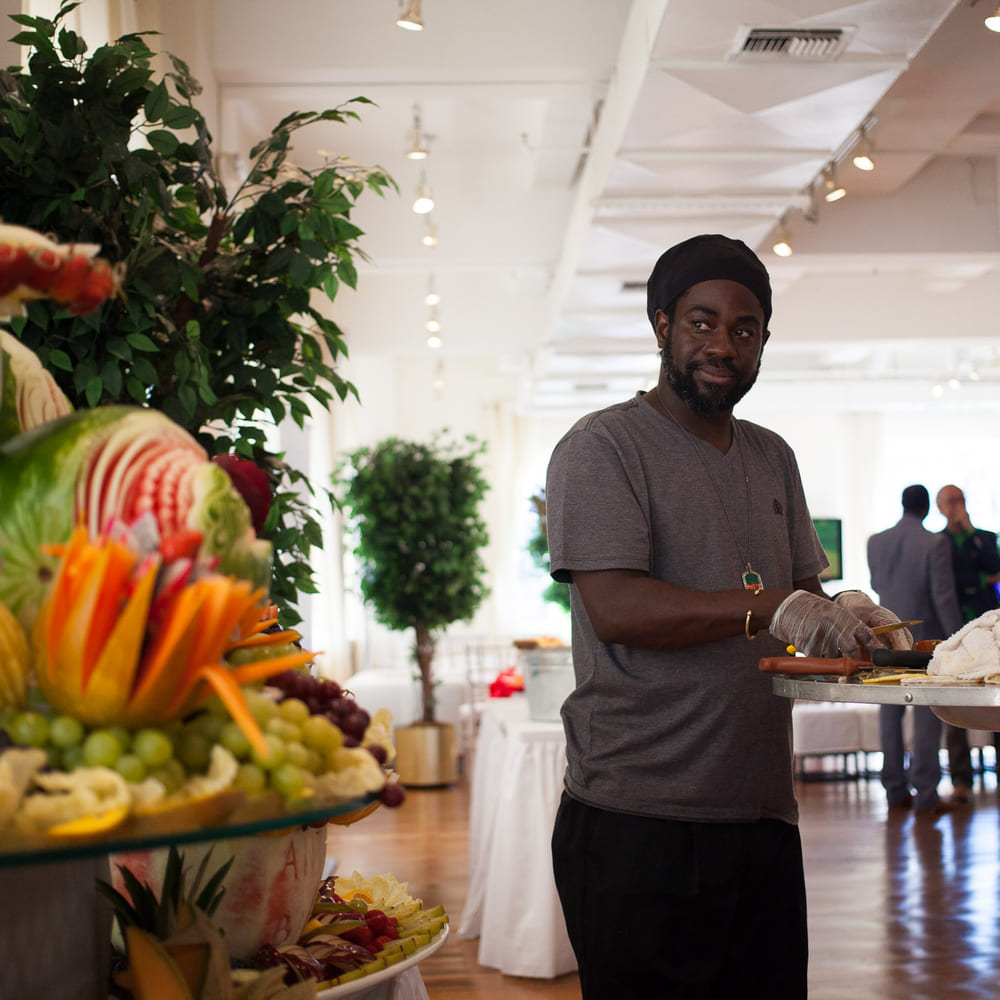 Chef Melvin Myers beside a tiered fruit display in a bright event space, carved melon and pineapple stacked behind him.