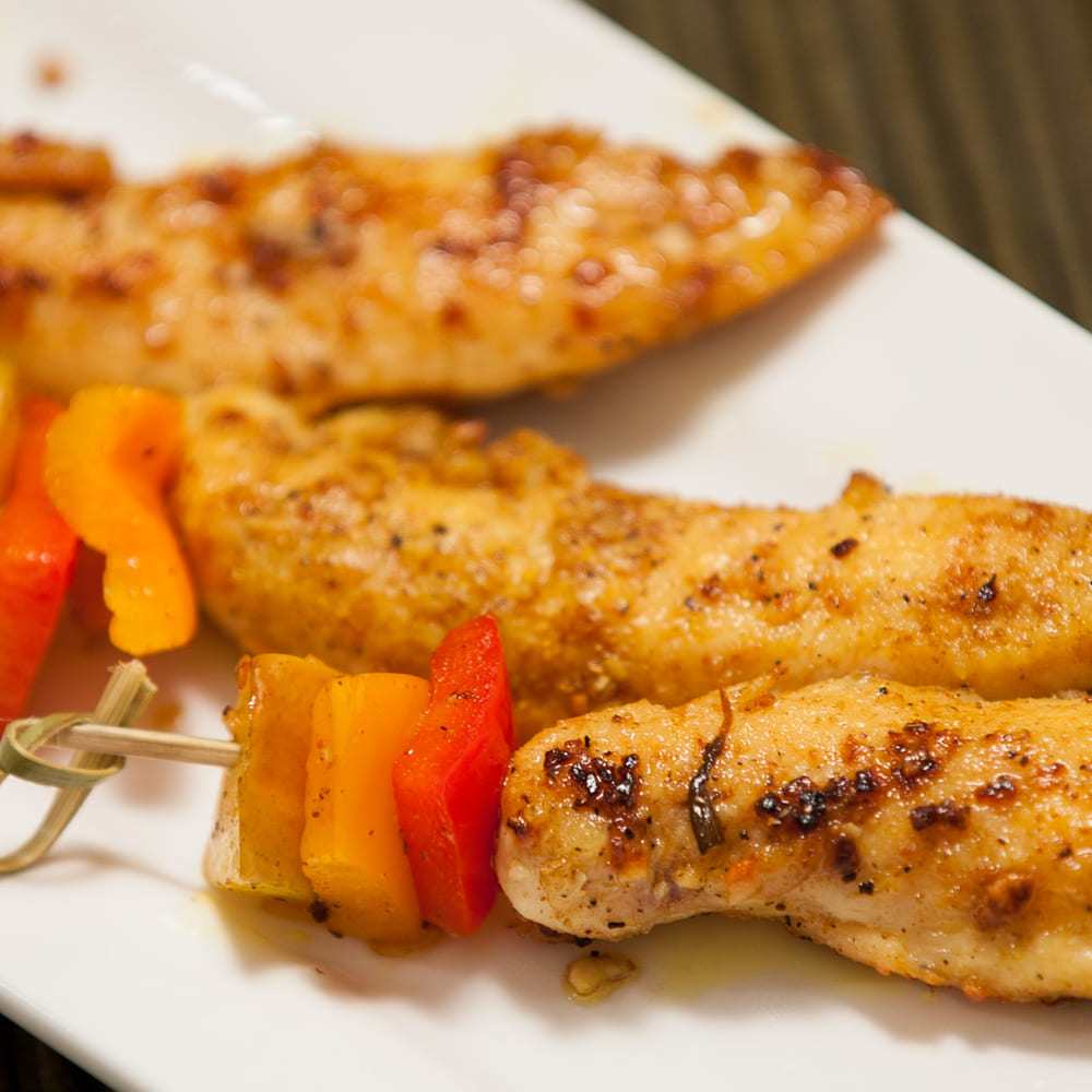 Three seasoned chicken tenders on a white plate with skewers of red and yellow pepper.