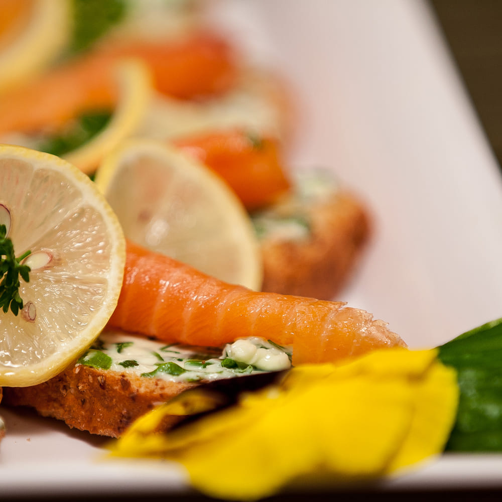 Smoked salmon canapés on seeded bread with herb butter, lemon slices and parsley on a white platter.
