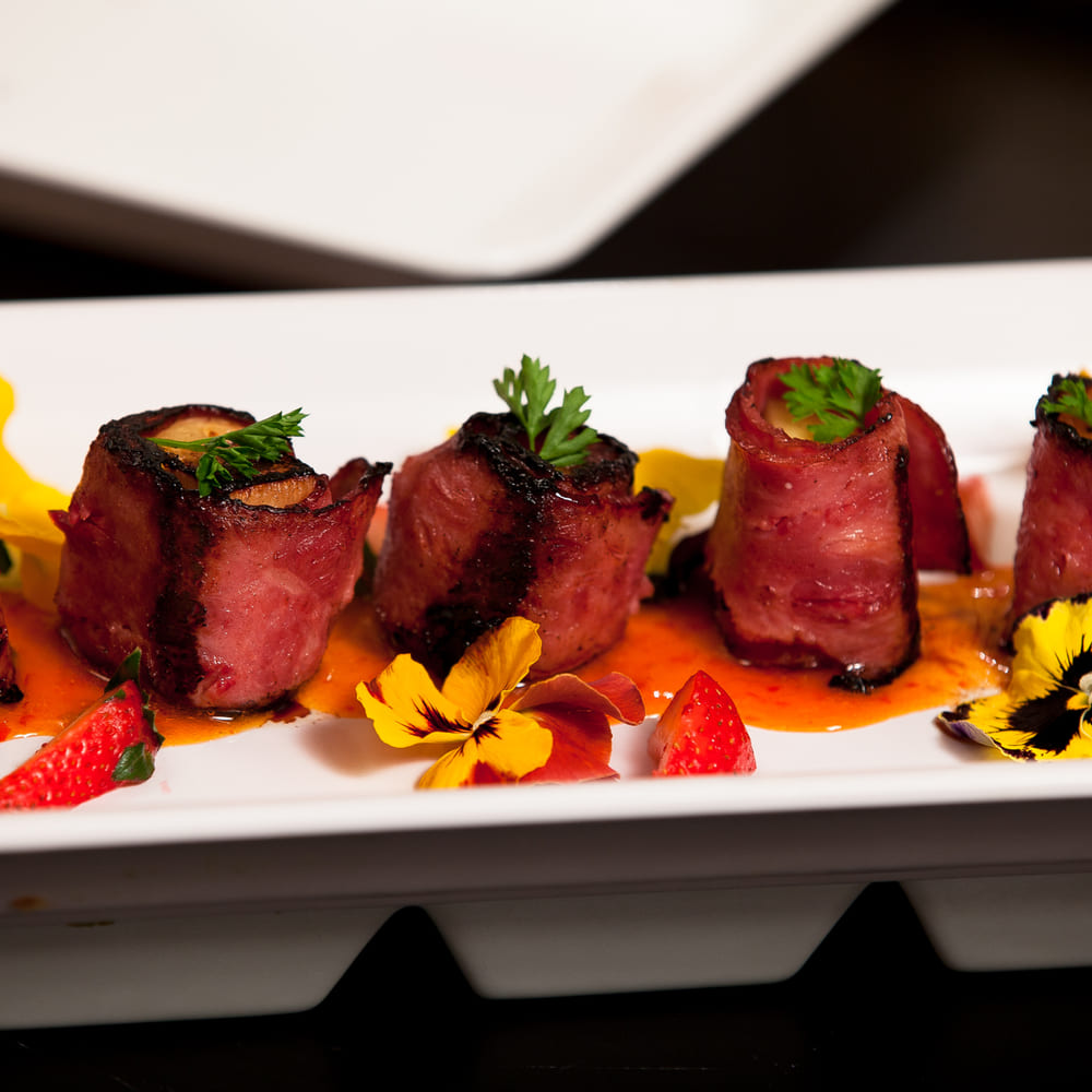 Bacon-wrapped medallions on a white plate with pansies, strawberries and a red pepper sauce.