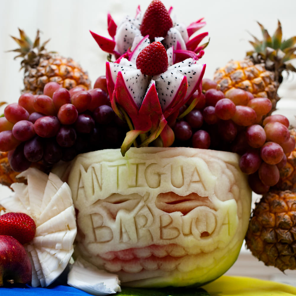 A watermelon carved with Antigua and Barbuda, topped with dragon fruit flowers and flanked by pineapples and grapes.