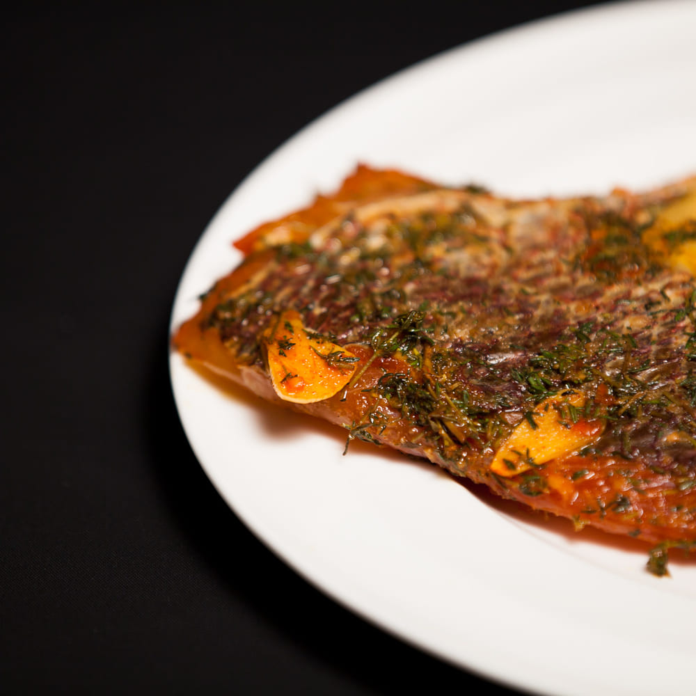 A whole roasted fish crusted with dill and garlic slices, served on a white oval plate against a black cloth.