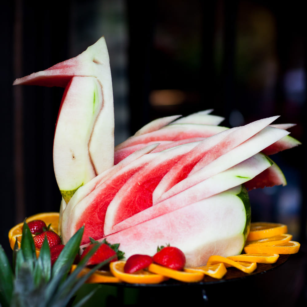 A swan carved from a whole watermelon, wings fanned in red and white, on a bed of orange slices and strawberries.
