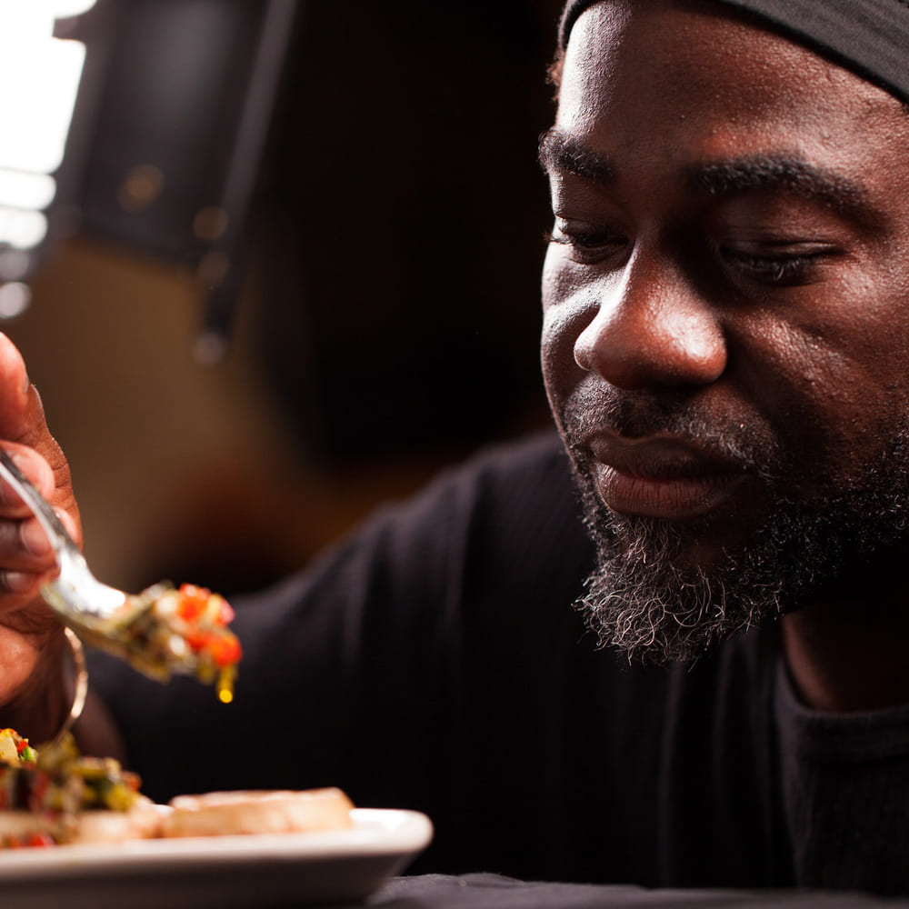 Chef Melvin Myers tasting from a fork over a plated dish, lit low against a dark kitchen.