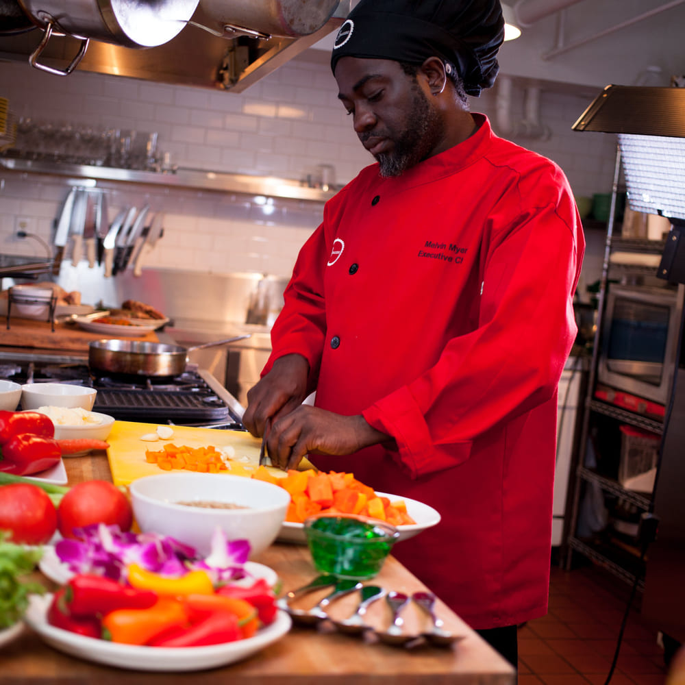 Chef Melvin Myers in a red red chef's coat dicing butternut squash at a professional kitchen station.