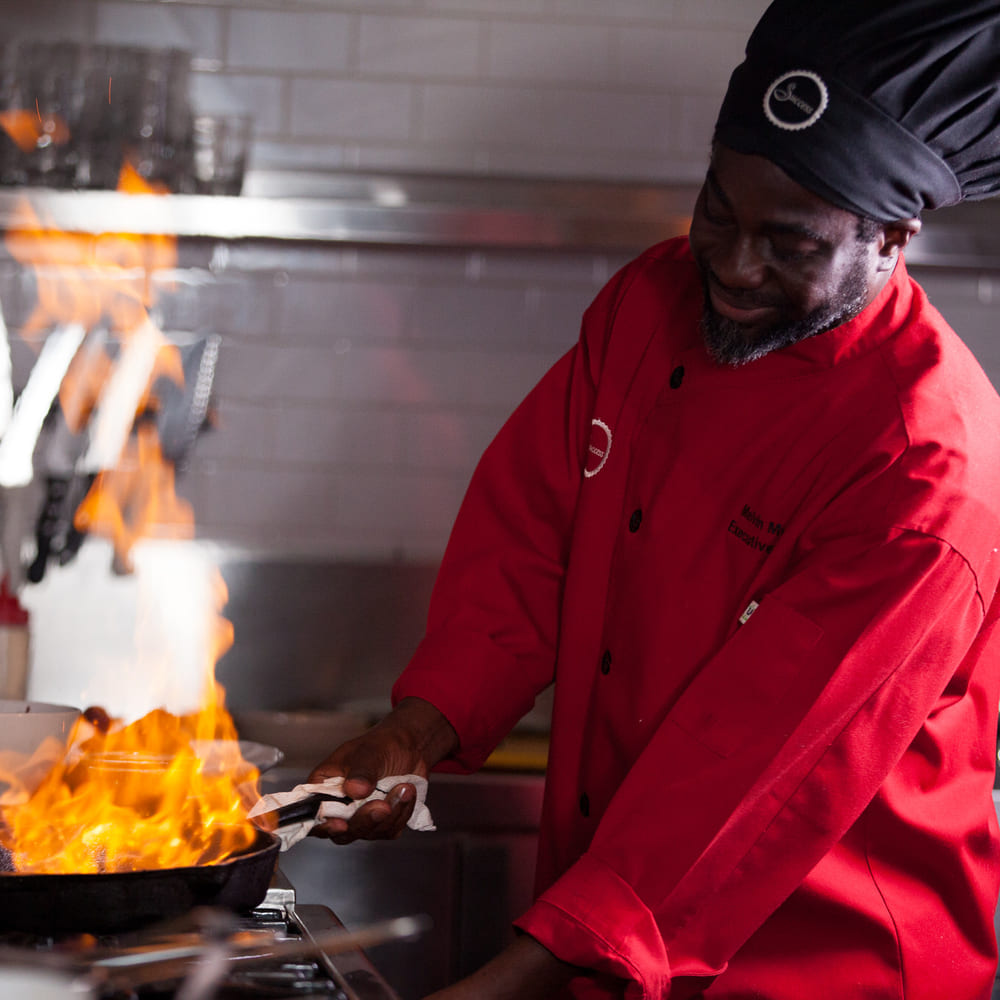 Chef Melvin Myers flambéing in a pan, flames rising, in a professional kitchen.