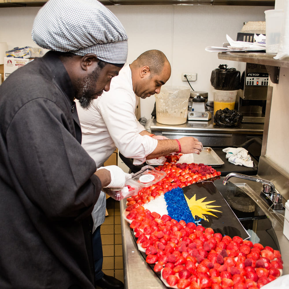Chef Melvin Myers and a second chef laying strawberries and blueberries into the Antigua and Barbuda flag on a kitchen tray.