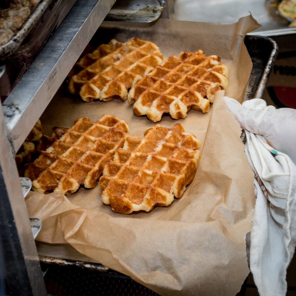 Freshly baked Liège waffles cooling on parchment in a commercial kitchen, a gloved hand steadying the tray.