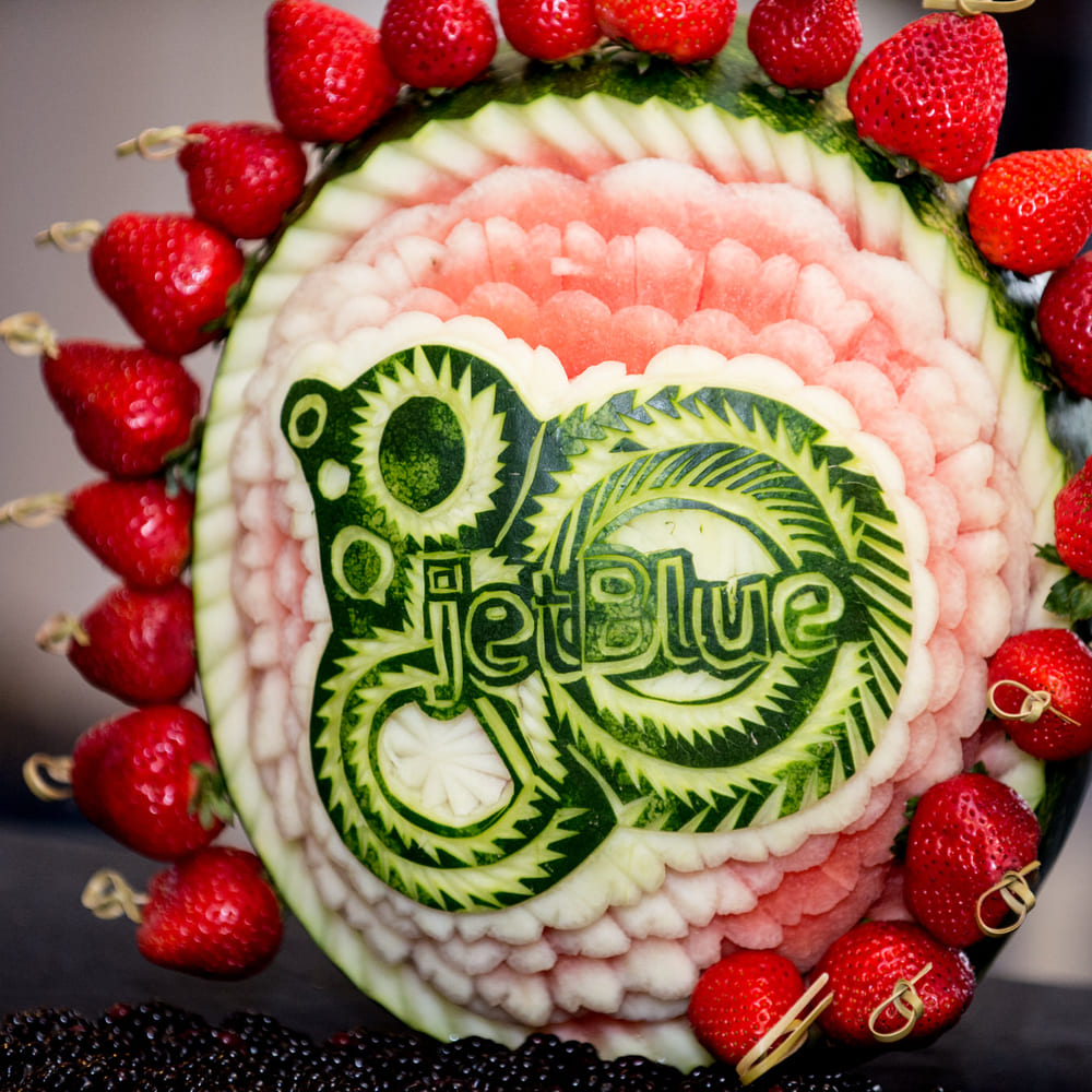 The JetBlue logo carved into a watermelon, surrounded by strawberries on skewers and blackberries.