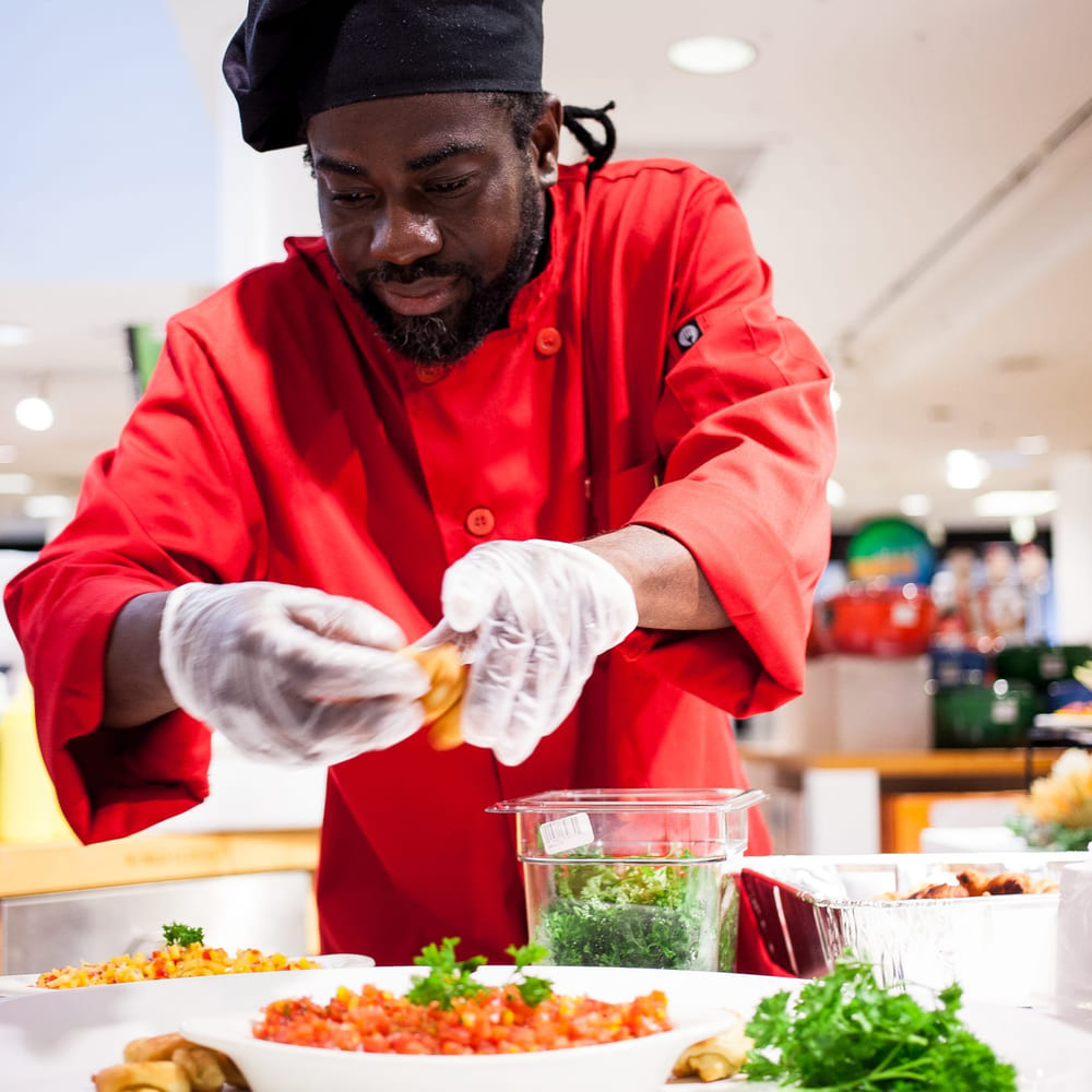 Chef Melvin Myers in a red chef coat plating at a department store food hall, a carved watermelon tower behind him.