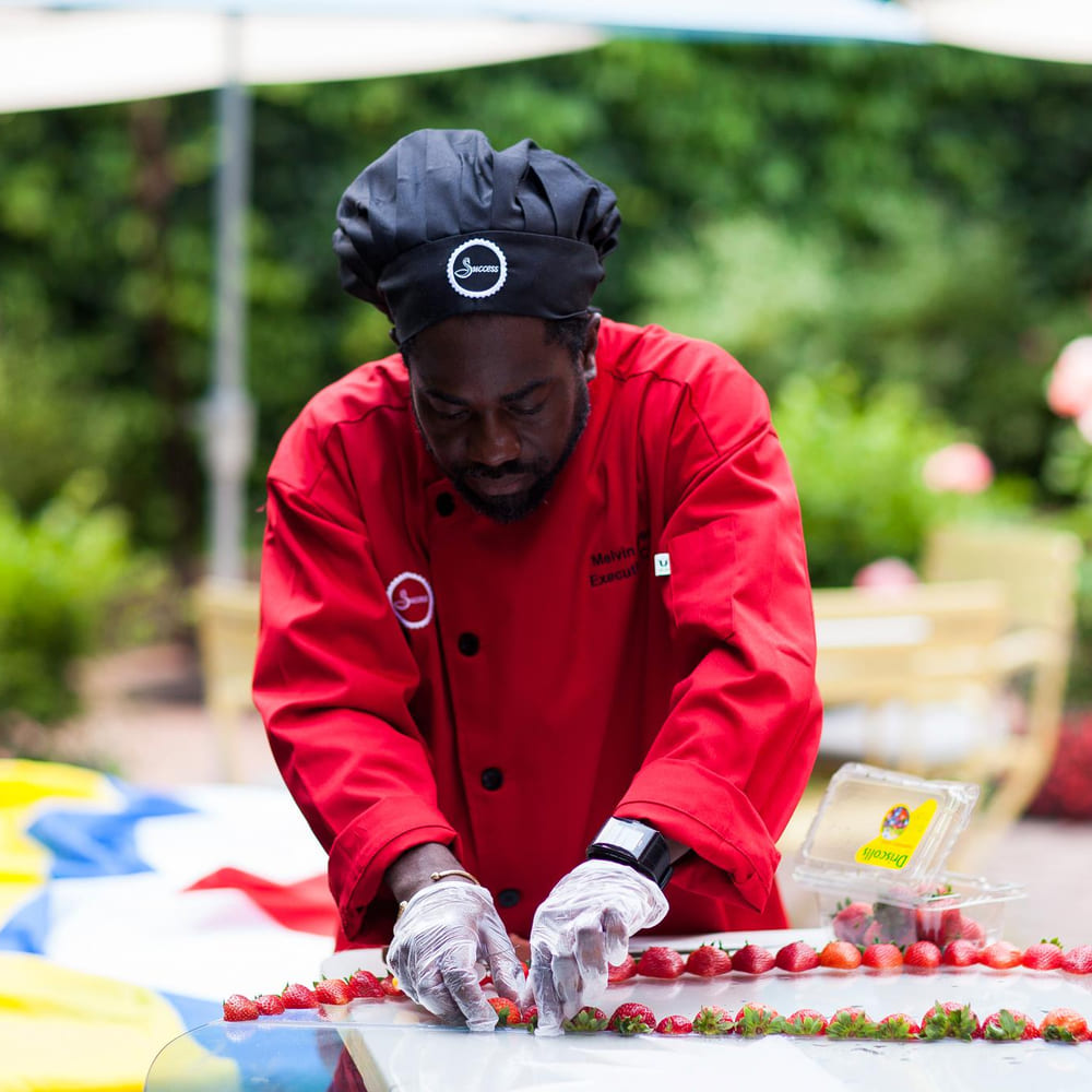 Chef Melvin Myers in a red Success Catering chef coat arranging strawberries at an outdoor event.