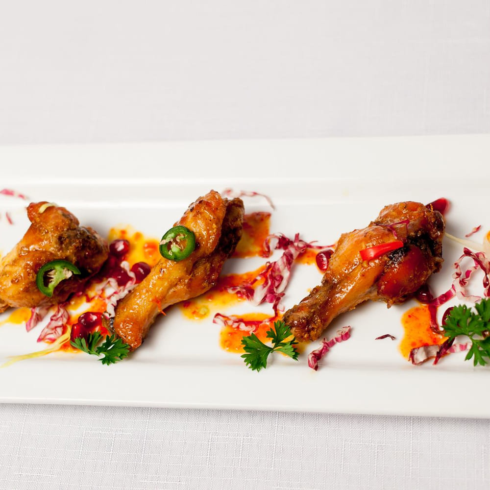 Four glazed chicken wings on a long white plate with jalapeño, pomegranate and a chilli drizzle.
